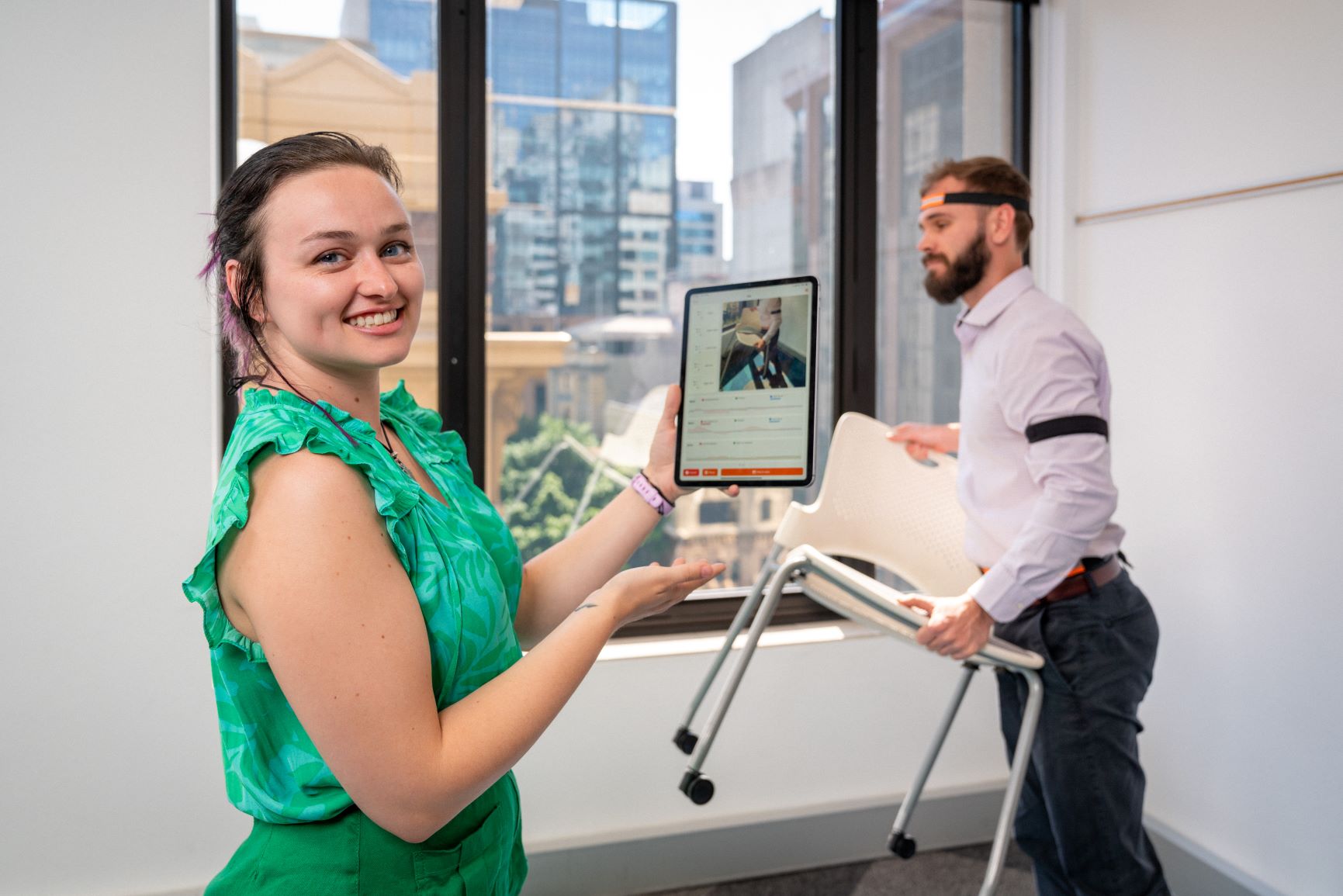 Smiling woman holds tablet while man carries chair.
