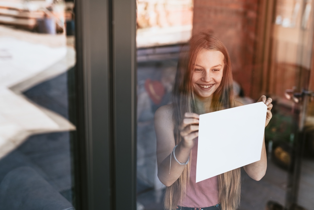 Girl smiling, holding blank white paper at window.